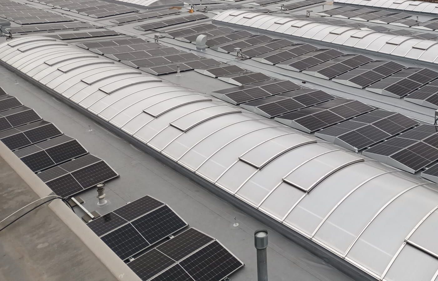 Dlouhý obloukový světlík na ploché střeše, kolem něj fotovoltaické panely a odvětrávací komínek